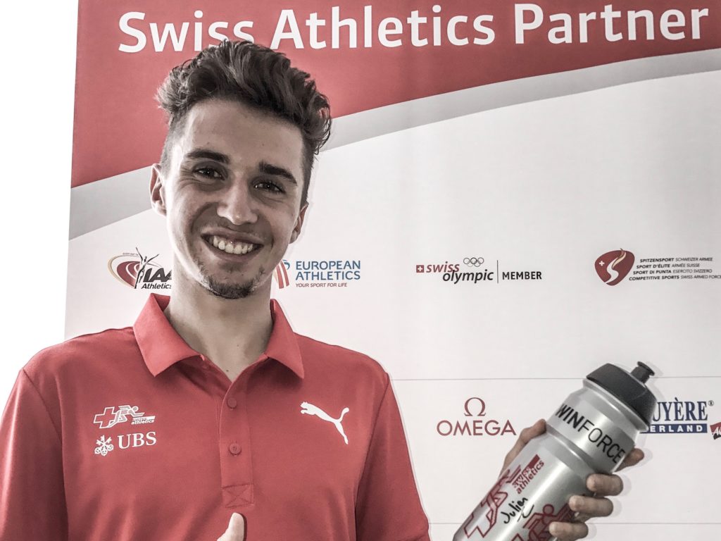 Julien Wanders posiert mit einer WINFORCE-Flasche vor der Sponsorenwand von Swiss Athletics